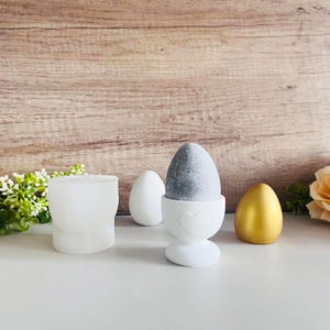 Könnte beinhalten: Drei Keramik-Eierbecher, einer mit einem grauen, gefleckten Ei darin, einer mit einem goldenen Ei darin und einer leer. Der Eierbecher mit dem grauen Ei hat ein Herzdesign auf der Vorderseite.