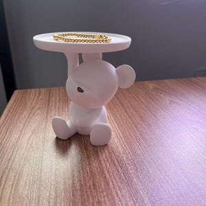 May include: White ceramic bear figurine holding a white tray with a gold beaded bracelet on top.
