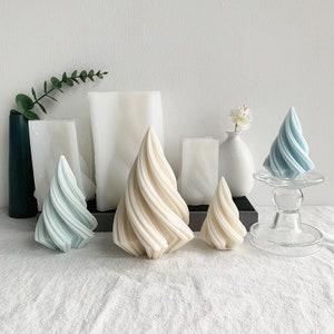 Può includere: Cinque candele decorative a forma di spirale, due sono blu chiaro e tre sono bianche. Le candele sono disposte su una superficie bianca con un piedistallo di vetro al centro.