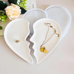 May include: Two white ceramic heart-shaped dishes, one with a gold necklace and the other with a gold ring. The dishes are designed to fit together like a puzzle.
