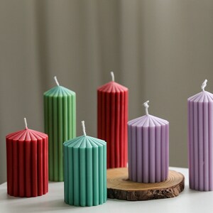 May include: Six pillar candles in various colors, including red, green, and purple. The candles have a fluted design and are arranged on a wooden surface.