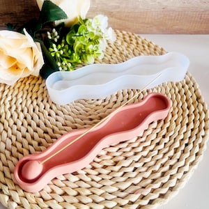 May include: A pink and white wavy incense holder with a gold incense stick. The holder is made of silicone and is designed to catch the ash from the incense stick.