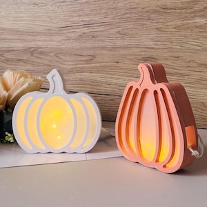 Könnte beinhalten: Zwei dekorative Kürbis-Lichter. Eines ist weiß, das andere korallenfarben. Beide strahlen ein warmes, gelbes Licht aus. Die Kürbisse aus Holz sind für Herbst- oder Halloween-Dekorationen gedacht.