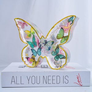 Könnte beinhalten: Eine transparente Schmetterlings-Skulptur aus Harz mit goldener Umrandung. Der Schmetterling ist mit bunten Schmetterlingen und Blumen gefüllt.