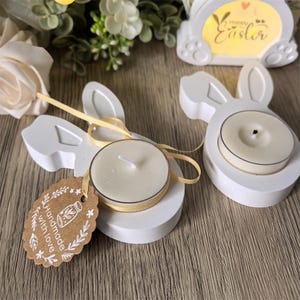 Może przedstawiać: Białe świeczniki w kształcie króliczka ze świeczkami typu tea light i żółtymi wstążkami. Etykieta głosi "Handmade with love". Z tyłu znajduje się znak "Happy Easter" z motywem królika. Tematem są Święta Wielkanocne.