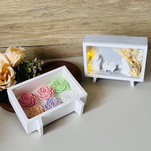 Könnte beinhalten: Eine weiße rechteckige Box mit sechs rosenförmigen Seifen in Rosa, Grün, Lila und Weiß. Daneben eine weiße Krippenszene mit warmem Licht im Inneren, die eine festliche Atmosphäre schafft.