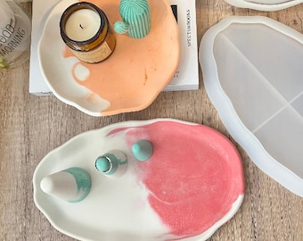 Può includere: Due vassoi ovali in resina. Un vassoio è bianco con un design marmorizzato rosa e presenta tre piccoli pezzi decorativi color turchese. L'altro vassoio è arancione con un design marmorizzato bianco e presenta un pezzo decorativo a forma di cactus color turchese e una piccola candela di vetro.