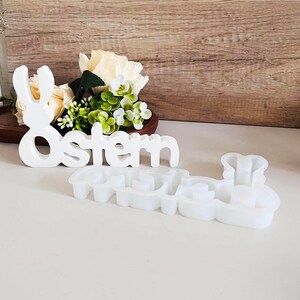 Op de afbeelding: Een wit houten bord met het woord "Ostern" en een konijnenuitsparing. Het bord ligt naast een witte siliconen mal van hetzelfde woord en dezelfde konijn.
