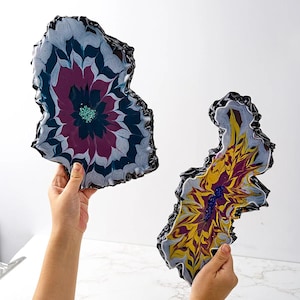 May include: Two resin coasters with a unique, abstract floral design. The first coaster features a dark purple and white flower with a green center. The second coaster features a yellow and white flower with a purple center.