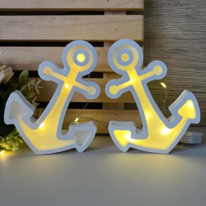 Op de afbeelding: Twee witte anker-vormige LED-lampen met gele verlichting. De nautische decoraties staan tegen een houten achtergrond. De lampen zijn verbonden door een reeks kleine lampjes.