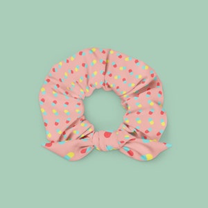 Puede incluir: Un scrunchie rosa con un patrón de píldoras amarillas, rojas y azules. El scrunchie está atado en un lazo.