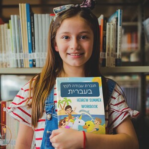 May include: A young person in a red and white striped shirt and blue overalls is holding a Hebrew summer workbook. The book has a colorful cover with a cartoon illustration of children playing on a beach with a unicorn and a duck.