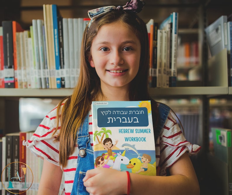 May include: A young person in a red and white striped shirt and blue overalls is holding a Hebrew summer workbook. The book has a colorful cover with a cartoon illustration of children playing on a beach with a unicorn and a duck.