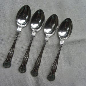 May include: Four silver spoons with decorative floral patterns on the handles. The spoons are arranged diagonally on a white, textured surface. The spoons have a shiny, reflective finish.