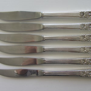 May include: A collection of five stainless steel dinner knives. Each knife has a curved blade and a handle with a ribbed design and floral detailing. The word "Community" is etched on the blade.