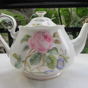 May include: A white porcelain teapot with a floral design featuring a pink rose and green leaves. The teapot has a gold trim around the rim and base.