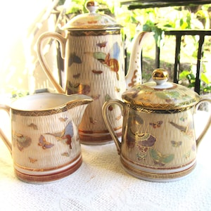 May include: A vintage porcelain tea set with a cream background and gold accents. The set includes a teapot, creamer and sugar bowl. The design features a delicate pattern of butterflies and flowers.