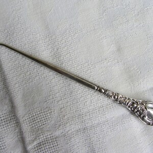 May include: A silver crochet hook with an ornate handle. The hook is long and thin with a small loop at the end. The handle is decorated with a floral design.