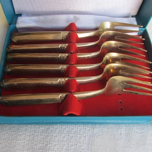 May include: A set of six gold-coloured forks presented in a blue box with a red velvet interior. Each fork is held in place by a red velvet holder. The box is open, displaying the silverware.