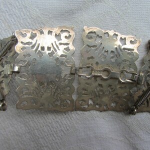 May include: A silver belt featuring a decorative, intricate design. The belt is made up of multiple connected pieces, each with a unique pattern. The belt is flexible and can be adjusted to fit different sizes.
