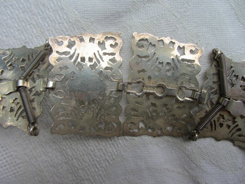 May include: A silver belt featuring a decorative, intricate design. The belt is made up of multiple connected pieces, each with a unique pattern. The belt is flexible and can be adjusted to fit different sizes.