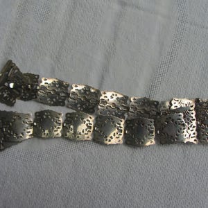 May include: A silver chain belt with a decorative, intricate design. The belt is made of linked, square-shaped pieces with a cutout pattern.