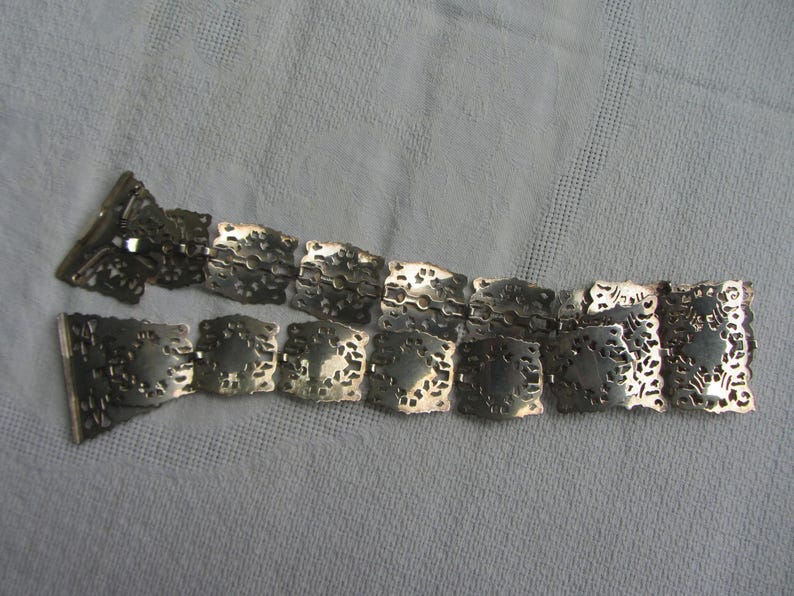 May include: A silver chain belt with a decorative, intricate design. The belt is made of linked, square-shaped pieces with a cutout pattern.