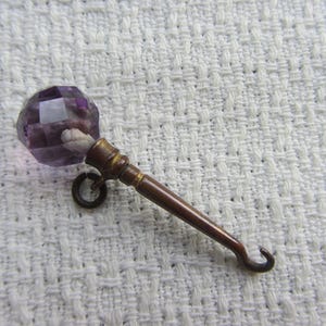 May include: A metal button hook featuring a faceted purple glass bead at the top. The handle is made of a tarnished bronze-coloured metal and includes a small circular loop near the top, ending in a curved hook designed for fastening buttons.