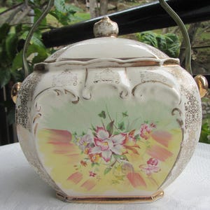 May include: A vintage ceramic biscuit jar with a lid and handle. The jar is cream-coloured with gold trim and a floral design in the centre. The floral design features pink and white flowers with yellow and green accents.