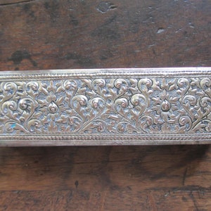 May include: A silver rectangular box with an intricate floral design. The box has a raised border and is decorated with a repeating pattern of leaves and flowers.