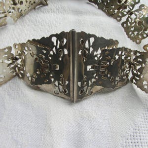 May include: A silver filigree belt with a decorative clasp. The belt is made of interlocking metal pieces that form a delicate pattern.