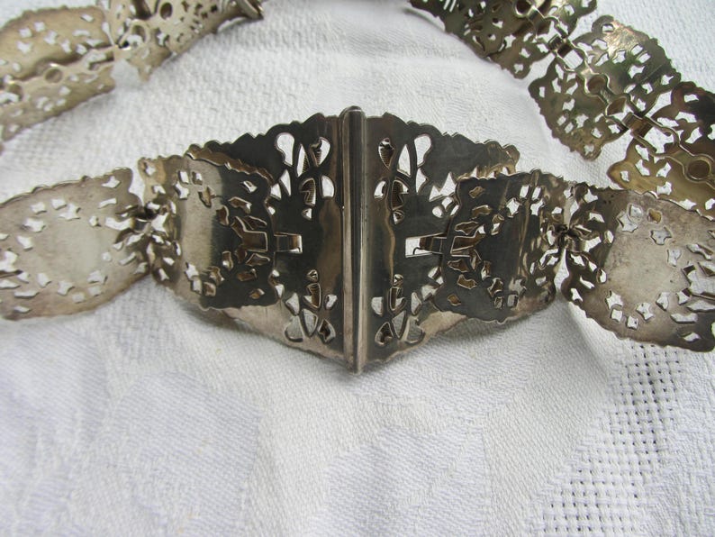 May include: A silver filigree belt with a decorative clasp. The belt is made of interlocking metal pieces that form a delicate pattern.