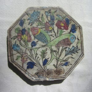May include: An octagonal ceramic tile with a hand-painted design. The tile features two birds surrounded by colourful flowers in shades of blue, pink, yellow, and red. The birds have blue and green bodies. The tile has a cracked, aged appearance.