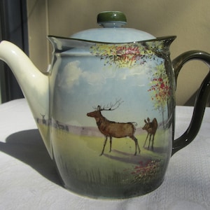 May include: A decorative ceramic teapot with a painted landscape scene. The teapot features a light blue body and lid, with a cream spout and dark green handle. The scene depicts deer in a field.