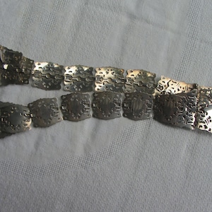 May include: A silver chain belt with a decorative, intricate design. The belt is made of linked, square-shaped pieces with cut-out patterns.