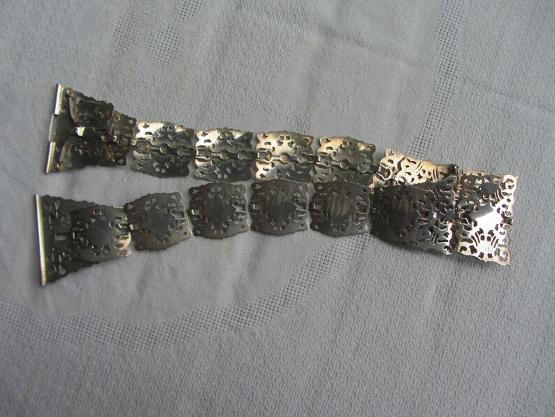 May include: A silver chain belt with a decorative, intricate design. The belt is made of linked, square-shaped pieces with cut-out patterns.