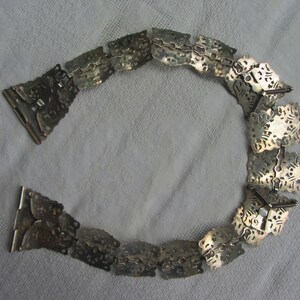 May include: A silver chain belt with intricate, ornate designs. The chain is made of linked, square-shaped pieces with a textured surface.