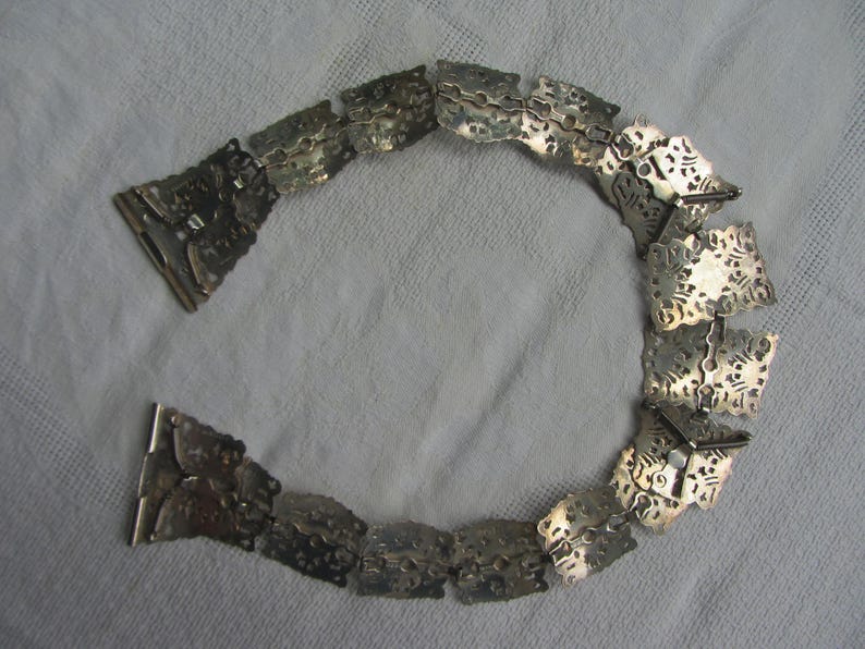 May include: A silver chain belt with intricate, ornate designs. The chain is made of linked, square-shaped pieces with a textured surface.