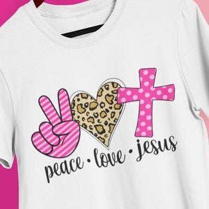 May include: White t-shirt with a pink peace sign, a pink and leopard print heart, and a pink cross with white polka dots. The text "peace. love. jesus" is printed below the design.