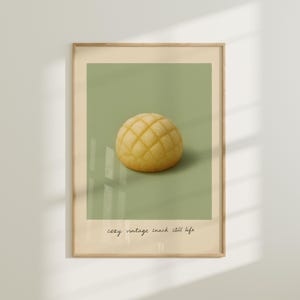 May include: A framed print featuring a close-up of a golden-yellow melon pan pastry against a muted green background. The print has the words "cozy vintage snack still life" in a handwritten font at the bottom.