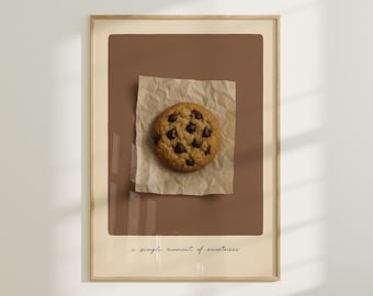May include: A framed print featuring a chocolate chip cookie on a piece of crumpled parchment paper. The cookie is round and golden brown with dark chocolate chips. The print has a brown background and the text "a simple moment of sweetness".