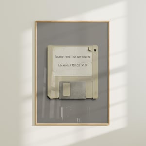 May include: Framed artwork featuring a vintage floppy disk with a handwritten label. The label reads "SOURCE CODE - DO NOT DELETE" and "LOCALHOST 127 OS V1.0". The artwork is set against a gray background within a wooden frame.