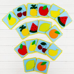Pode incluir: Um conjunto de 12 cartas de frutas de feltro correspondentes. Cada carta apresenta uma fruta diferente, incluindo abacaxi, maçã, banana, melancia, pêra, limão, laranja e cerejas. As cartas são azuis com bordas festonadas amarelas.
