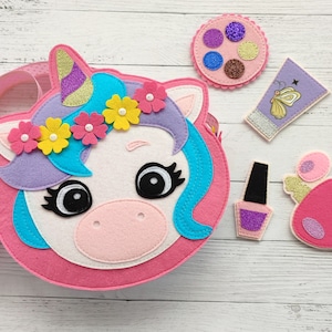 May include: A pink felt unicorn purse with a blue mane and a rainbow of flowers around its head. The purse comes with a set of felt accessories, including a makeup palette, a tube of lotion, a bottle of nail polish, and a perfume bottle.