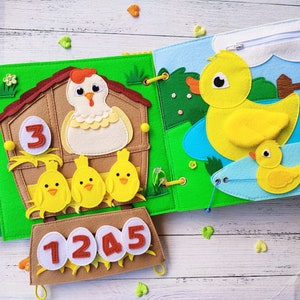 Pode incluir: Um livro de feltro para crian&ccedil;as com um pato amarelo, pintinhos e uma galinha em um galinheiro. O livro tem os n&uacute;meros 1, 2, 3, 4 e 5 na capa.