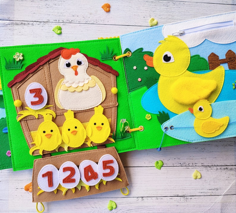 Pode incluir: Um livro de feltro para crian&ccedil;as com um pato amarelo, pintinhos e uma galinha em um galinheiro. O livro tem os n&uacute;meros 1, 2, 3, 4 e 5 na capa.