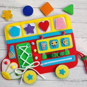 May include: A red felt bus toy with various activities for children. The bus has a zipper, laces to tie, a shape matching section, and a section with felt characters. The bus is decorated with yellow, blue, and green felt.