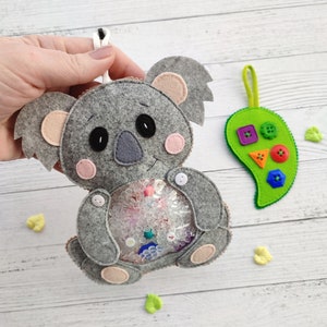 May include: A grey felt koala toy with pink ears and a clear plastic window in its belly filled with colourful beads. The koala is holding a small white button in its hand.