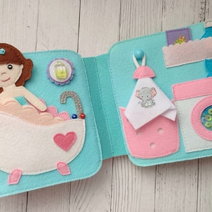 May include: A felt book with two pages. The left page shows a girl in a bathtub with bubbles and a bottle of soap. The right page shows a washing machine with a basket of laundry and a towel with an elephant on it.