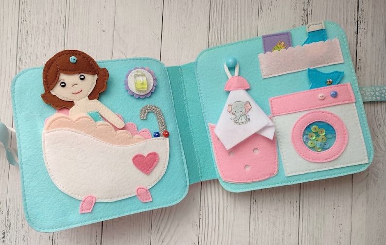 May include: A felt book with two pages. The left page shows a girl in a bathtub with bubbles and a bottle of soap. The right page shows a washing machine with a basket of laundry and a towel with an elephant on it.